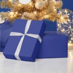 Blue holiday gift boxes with white ribbons under a decorated Christmas tree in a bright festive setting.