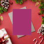 Stack of bright pink stationery cards beside notebook, pencil, and green plant on wooden surface.