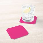 Glass of ice and lime on a bright pink coaster with a second coaster on light wood table.