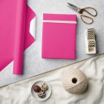 Hot pink wrapping paper with wrapped gift, gold scissors, twine, chocolates, and tape on neutral background.