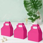 Three magenta cardboard favor boxes with handles beside Hershey’s Kisses and green plant on white tabletop.