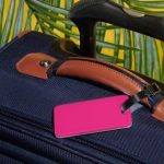 Pink luggage tag on navy suitcase handle with brown accents, framed by tropical leaves and yellow background.
