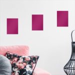 Three magenta rectangle art prints above pink floral cushion couch with black wireframe chair in modern interior.